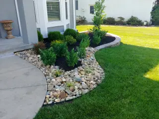Curved front foundation bed with river rock, evergreen shrubs, perennials, and dark mulch beside a manicured lawn